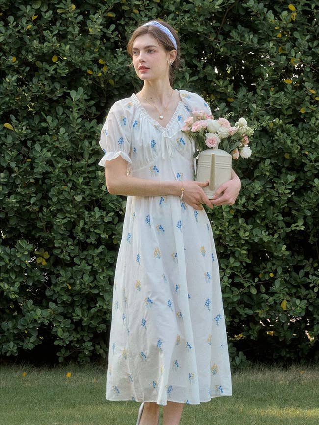 Person in a vintage-style dress holding a vase of flowers, standing in front of greenery