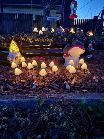 Garden furniture  decorated with assorted  mushroom-shaped lights and ornaments, including a butterfly and ample  mushroom figurines among flowering plants and mulch
