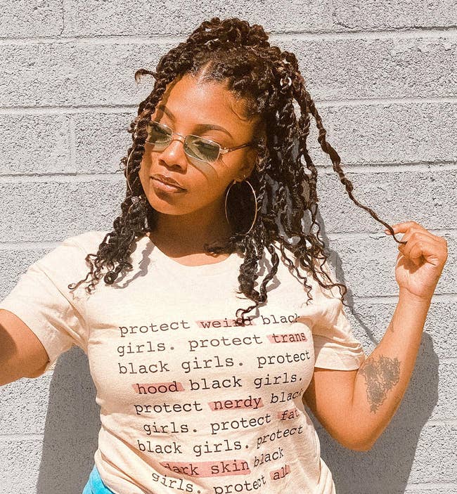 model in protect black woman tee that says protect weird black girls, protect trans black girls, protect hood black girls, protect nerdy black girls, protect fat black girls, protect dark skin black girls, protect all black girls