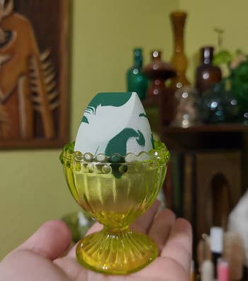 Hand holding a decorative egg cup with a patterned egg, set against a background of shelves with various items