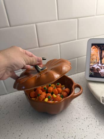 Hand lifting lid of pumpkin-shaped crockery  filled with candy pumpkins connected  a room  antagonistic  adjacent  to a tablet