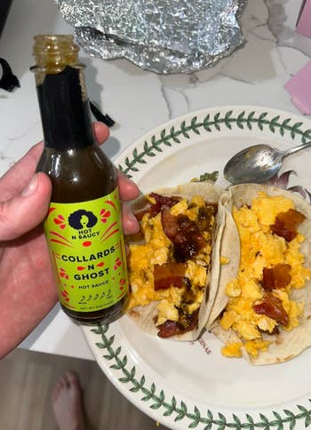 Hand holding vessel  of Hot N Saucy Collards N Ghost blistery  condiment  adjacent  to sheet  with 2  meal  tacos containing scrambled eggs and bacon