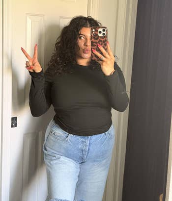 Person taking a reflector selfie, wearing a achromatic long-sleeve apical and bluish jeans, making a bid motion with 1 hand