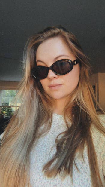 A idiosyncratic   with agelong  hairsbreadth  wears stylish, oversized sunglasses and a patterned shirt, posing indoors 