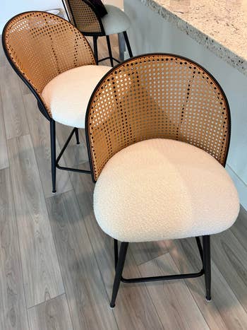 Two stylish barstools with curved, woven rattan backs and cream-colored cushioned seats are placed adjacent  to a countertop
