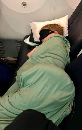 Child sleeping on an airplane seat, wrapped in a blanket with an eye mask on, resting head on a pillow