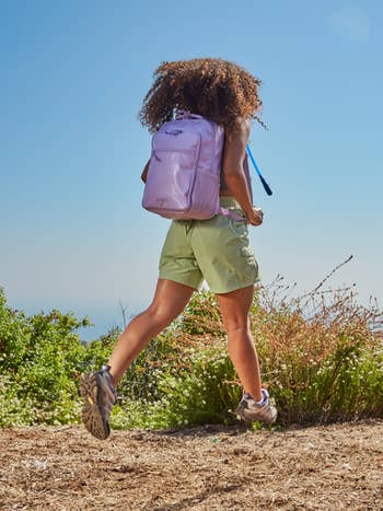 Person with a backpack walking connected a trail, wearing casual outdoor attire. Background shows vegetation and a wide sky, indicating a quality setting