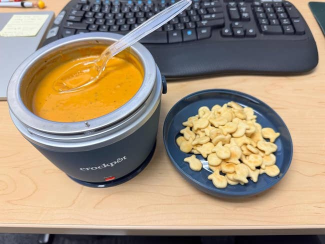 Sup di crockpot dengan sendok di sebelah sepiring kerupuk berbentuk ikan di atas meja dengan keyboard di dekatnya