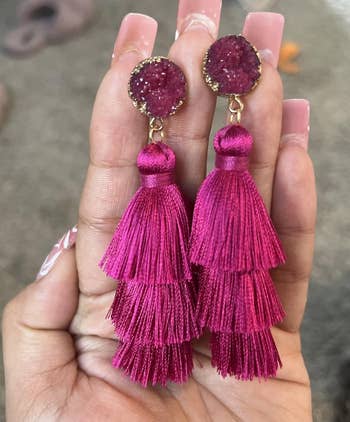 Hand holding pink tassel earrings with sparkling round studs, shown indoors on a carpeted floor with slippers in the background