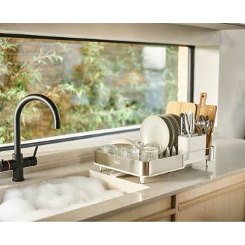 Kitchen sink with foam, a black faucet, and a dish rack holding plates, glasses, and utensils next to a large window