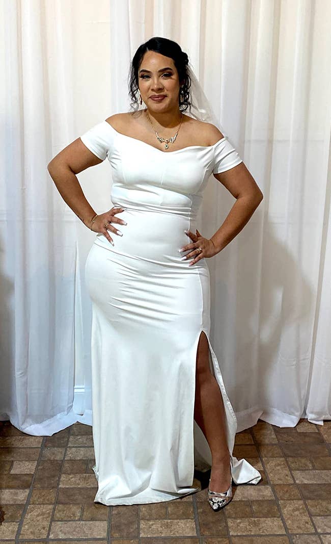 Reviewer wearing floor-length off the shoulder white dress with side leg slit and diamond necklace in front of a white curtain