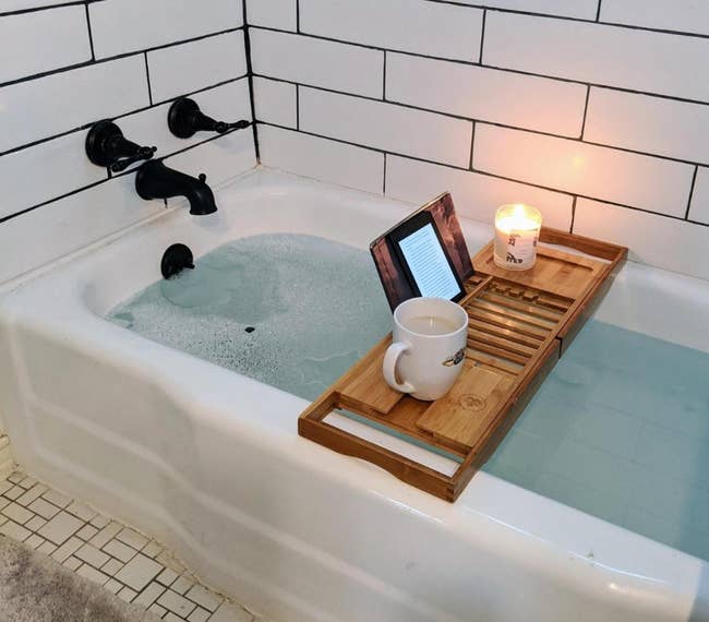 kindle on wooden tray over a filled bathtub, with a lit candle and a mug beside it