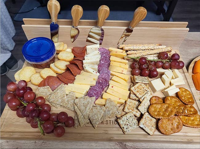 A light wood cheese board with crackers and cheese on it, plus a back board with four cheese knives attached 