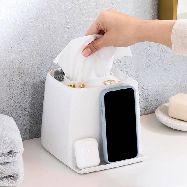 Hand reaching for tissues from a multi-functional organizer, with a phone and earbuds docked, on a bathroom counter