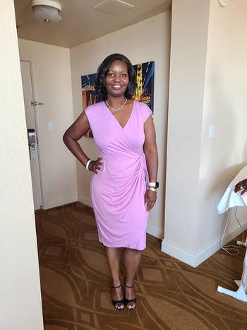 Person in a knee-length wrap dress, standing in a room, smiling with hands on hips