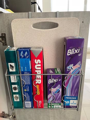 Organized cabinet door storage with various kitchen wrap boxes, including aluminum foil and plastic wrap, neatly arranged in a metal rack