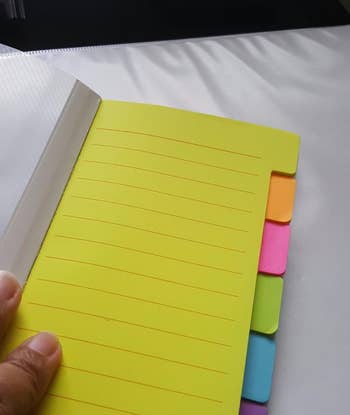 A hand holds a yellow notebook with colorful sticky tabs 