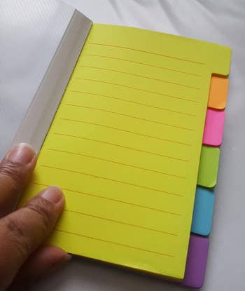 A hand holds a yellow notebook with colorful sticky tabs 