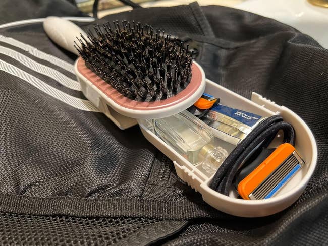 A hairbrush with a storage compartment containing toiletries such as a razor and a small bottle, shown resting on a travel bag