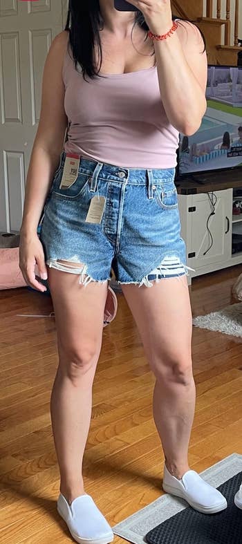 Person wearing a pink tank top, distressed denim shorts, and white slip-on shoes, standing indoors, with price tags visible on the shorts