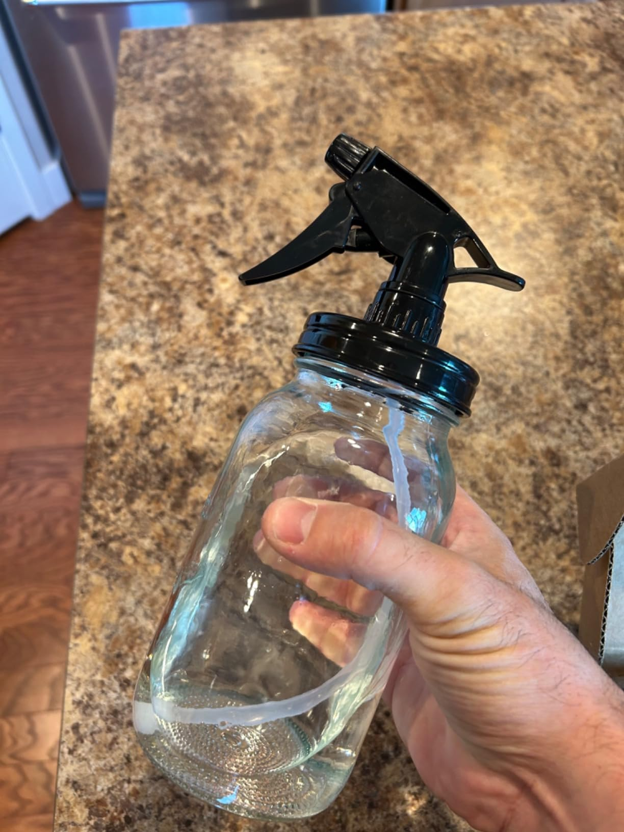 A manus  holding a transparent spray vessel  with a achromatic  nozzle, connected  a room  countertop
