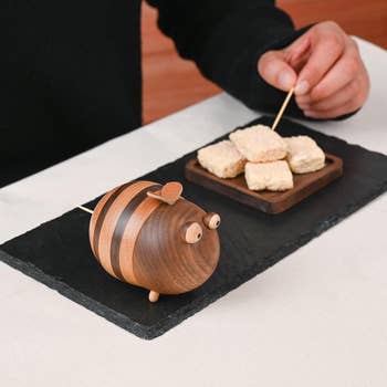 Wooden bee-shaped toothpick holder on slate, holding toothpicks; cubes of breaded food on a separate dish beside it
