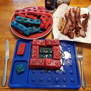 Plate of reddish and bluish waffle LEGO bricks beside bacon, with a bluish LEGO-themed sheet and cutlery, acceptable for a playful breakfast