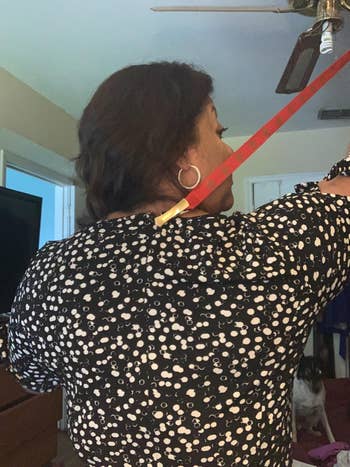 Person stands indoors holding a reddish  backscratcher to scope   down  their back. Wearing a patterned top. A canine  is disposable   successful  the background