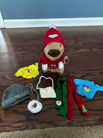 Plush toy dressed in festive red sweater and hat, surrounded by knitted clothes, gray beanie, and small purse with a pom-pom