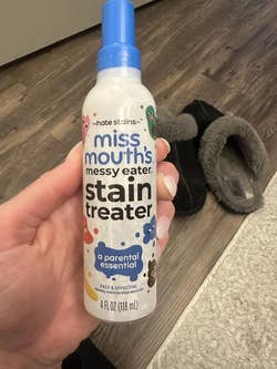Hand holding a bottle of Miss Mouth's Messy Eater Stain Treater near slippers. The product is a stain remover for parents