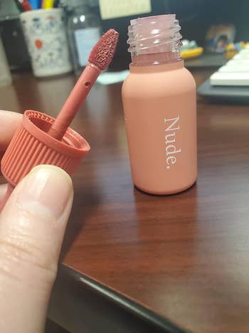 Open nude lipstick bottle with applicator held nearby, on a desk with various office items in the background