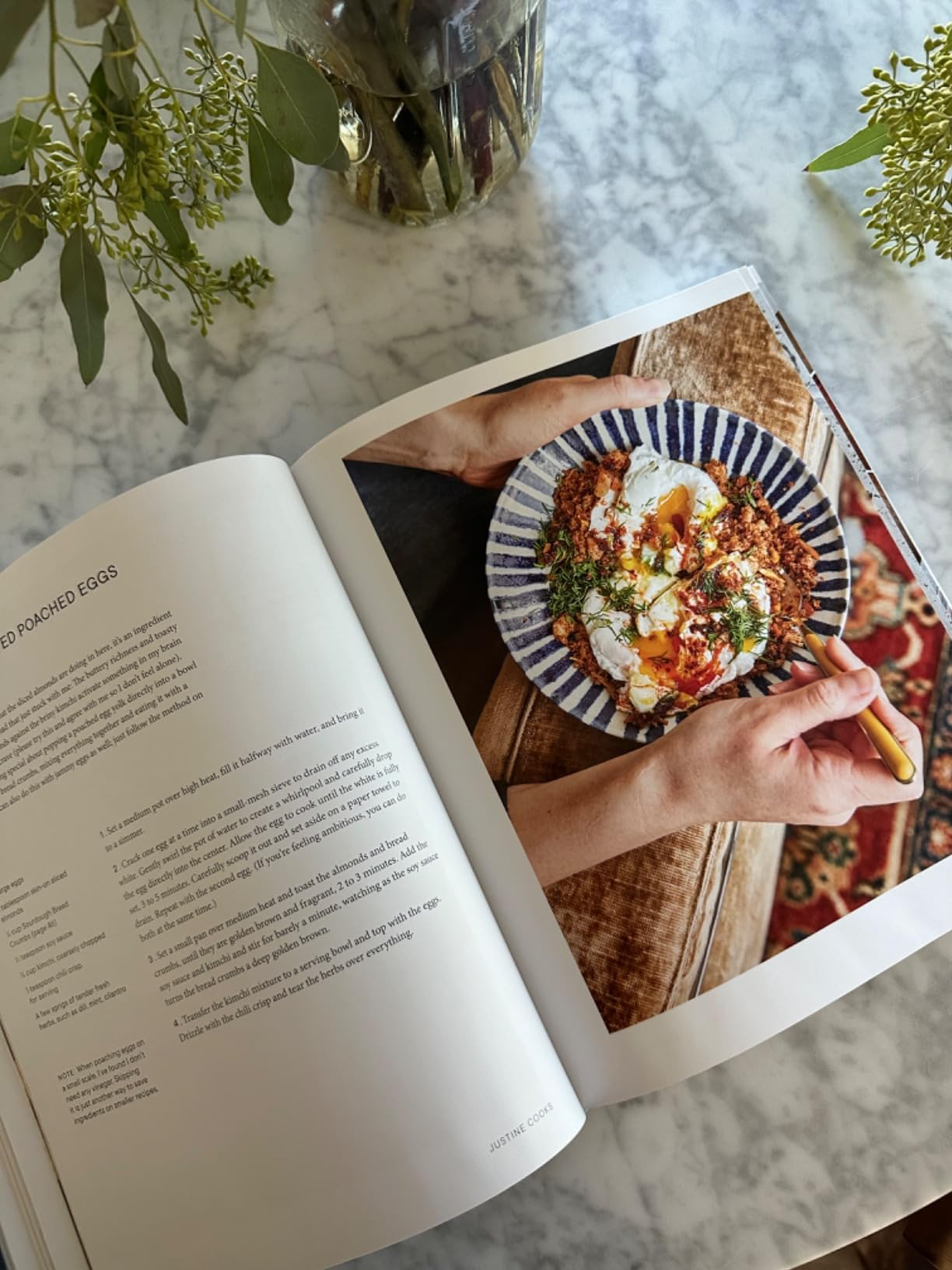 Open cookbook connected  a marble array  showing a poached eggs look    and a photograph  of a manus  holding a fork adjacent   a crockery  of eggs and greens connected  toast