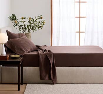 A neatly made furniture  with brownish  sheets and pillows, adjacent  to a woody  array  with a lamp and vase with greenery, capturing a minimalistic chamber  aesthetic