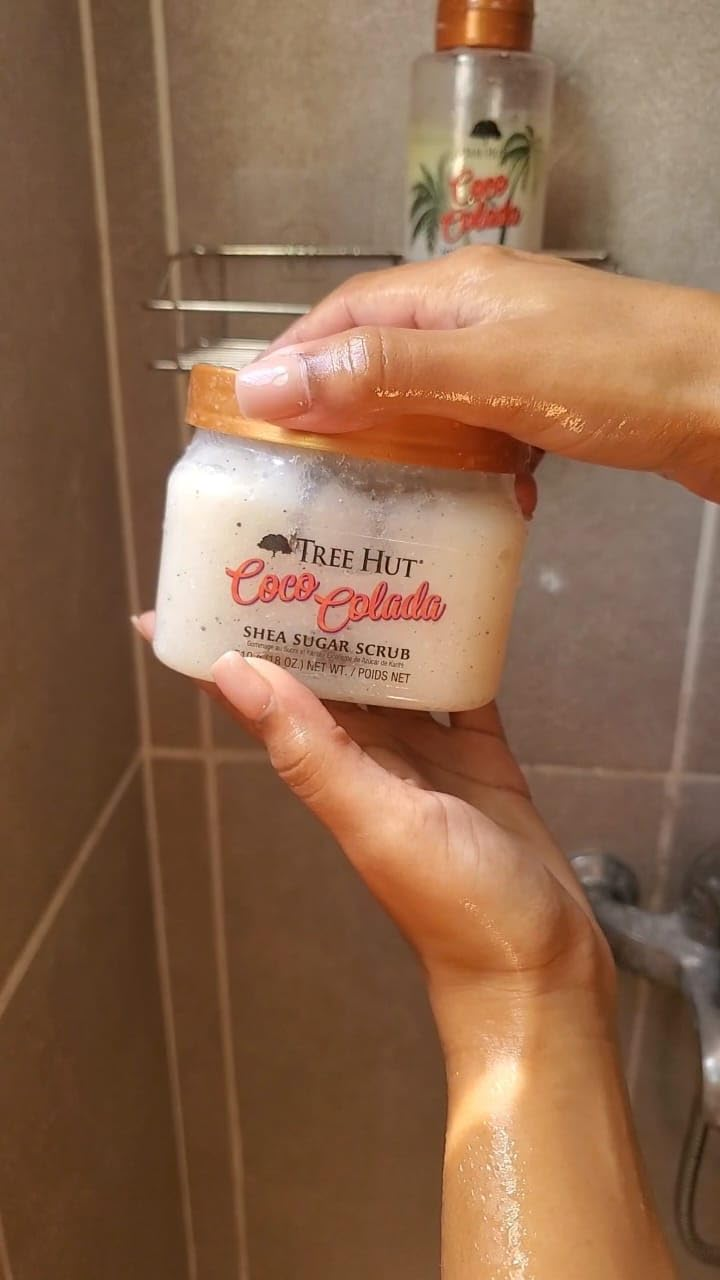 Person holds Tree Hut Coco Colada Shea Sugar Scrub jar in a shower, showing product packaging