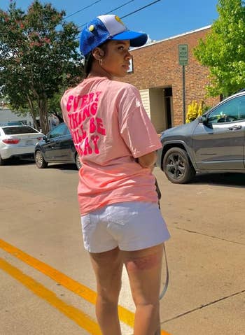 Person standing on a street wearing a pink t-shirt, shorts, a cap, and sneakers