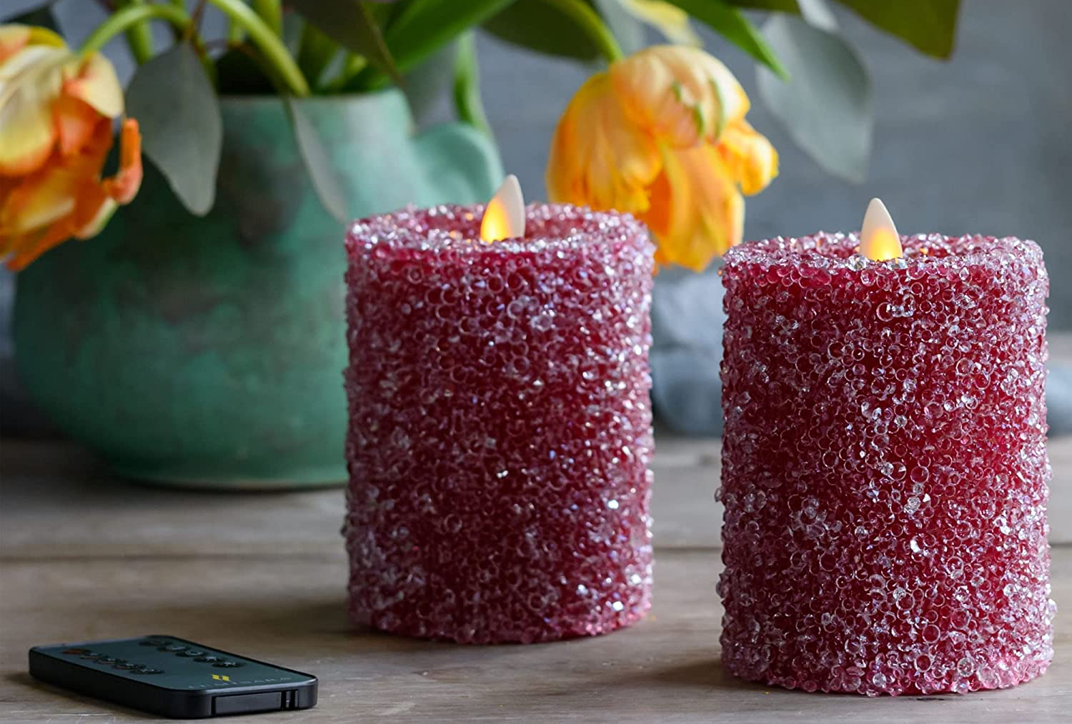 Two red crystal beaded flameless candles on top of wooden table next to small black remote