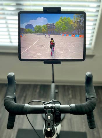 A stationary bike setup with a tablet attached, displaying a virtual cycling simulation with several cyclists riding on a paved path in a park setting