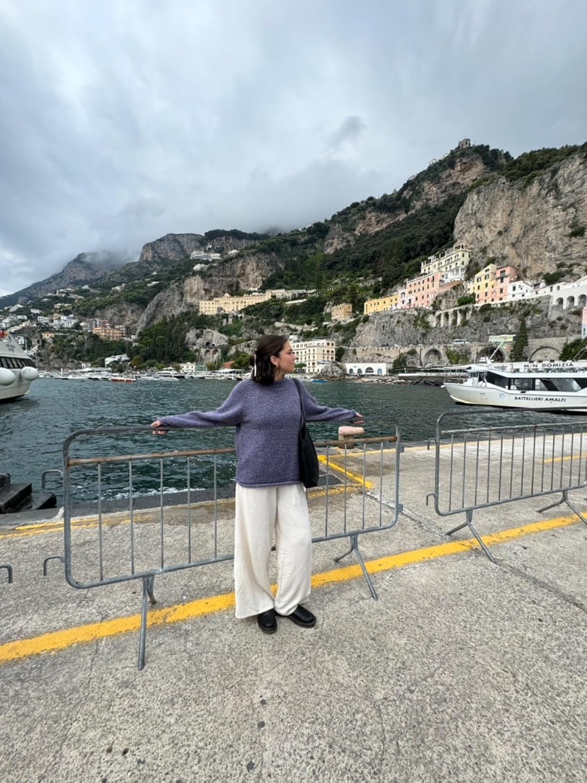 Person stands by a harbor with scenic hills and boats, wearing a cozy sweater and wide pants, holding a black bag, suggesting a relaxed travel style