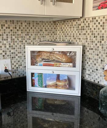Bread container  connected  a room  antagonistic  with a loaf of breadstuff  and hamburger buns stored inside. Mosaic tile backsplash is visible