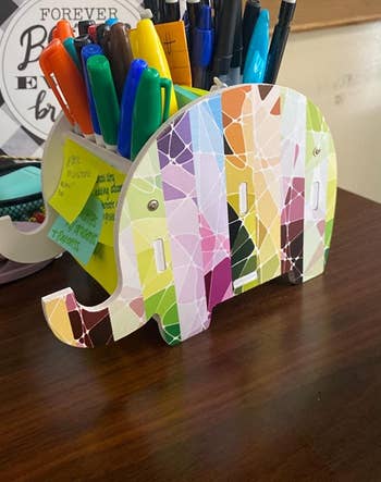 the colorful elephant holder holding a bunch of pencils and sharpies, with sticky notes on the side