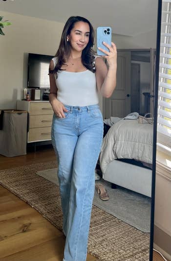Person takes a reflector selfie successful a modern bedroom, wearing a achromatic vessel apical and bluish jeans