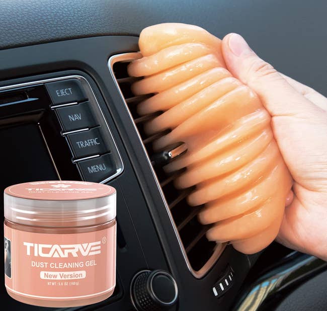 a hand holding the peach colored cleaning gel, which is being used to clean a car vent, with a tub of the cleaning gel in the bottom corner
