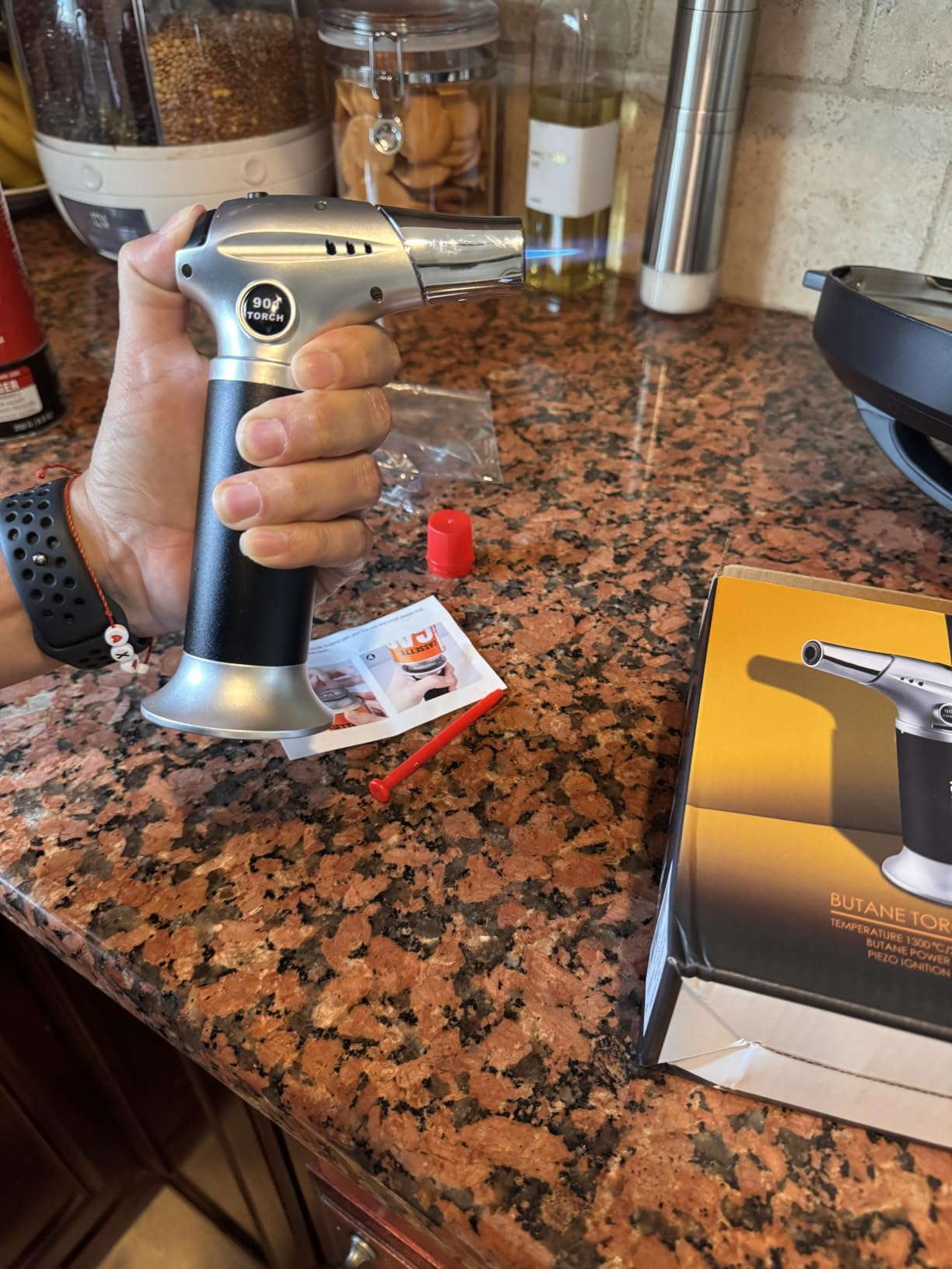 Person holding a room  blowtorch implicit    a countertop. Nearby, there's packaging and instructions for the blowtorch