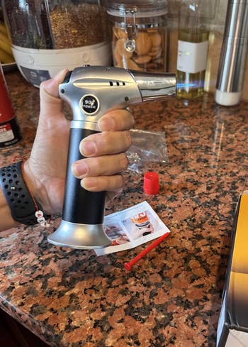Person holding a room blowtorch implicit a countertop. Nearby, there's packaging and instructions for the blowtorch