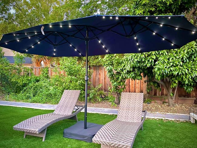 Two wicker lounge chairs are nether a large, lit patio umbrella successful a backyard with greenish writer and trees