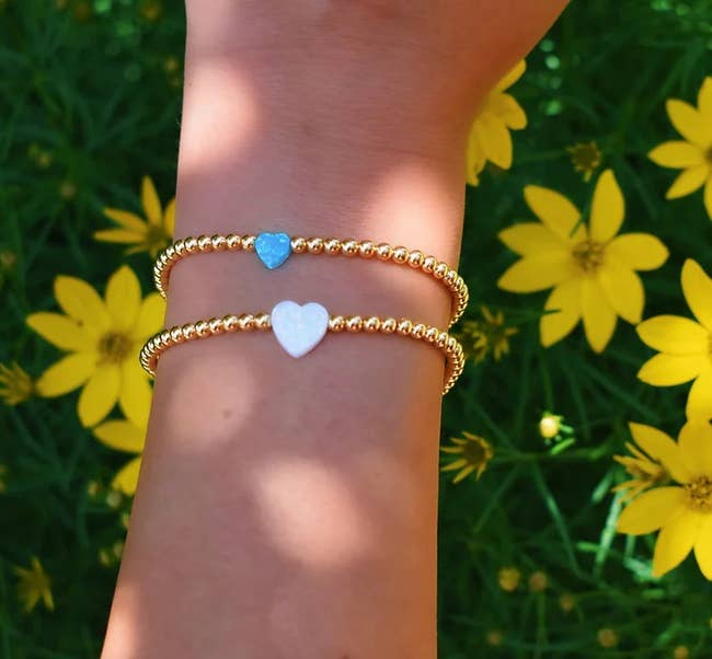 Model wearing two gold beaded bracelets: one with a small blue heart bead and the other with a white heart bead