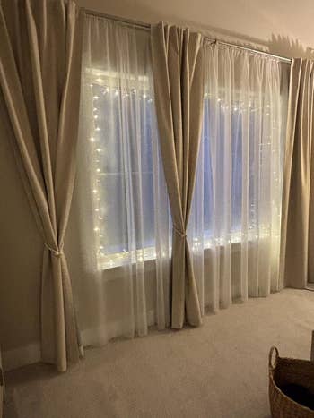 Large windows with sheer and dense  curtains, adorned with drawstring  lights, beside a woven handbasket  connected  the carpeted floor