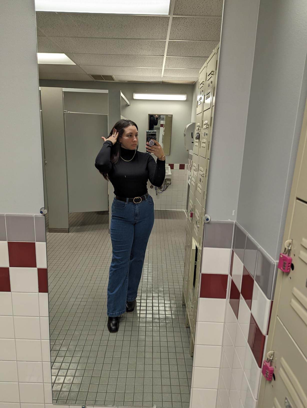 Person wearing a achromatic  apical  and high-waisted jeans, taking a reflector  selfie successful  a locker room
