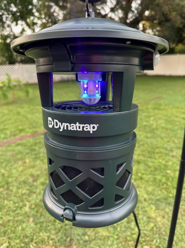 Dynatrap insect trap hanging outdoors, featuring a protective grill plan  and emitting a subtle bluish  glow