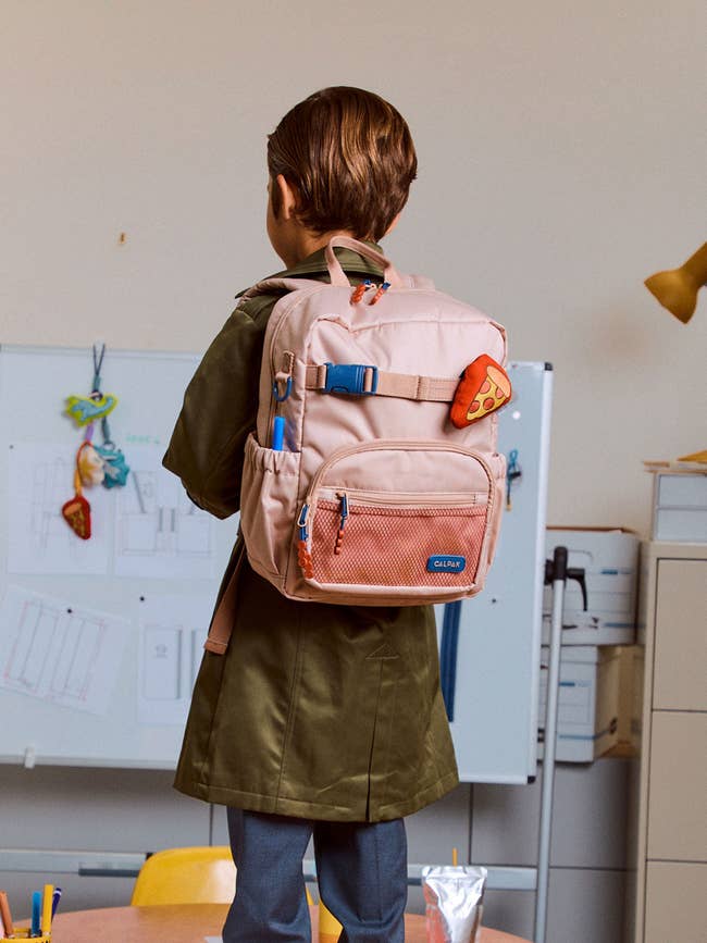 Child with a airy backpack featuring a pizza portion keychain stands successful a schoolroom with creation connected the whiteboard, suggesting back-to-school shopping